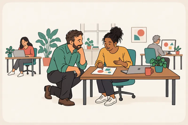 A manager kneels beside a team member at her desk and listens as she explains a sketch on a page, while two colleagues work in the background.